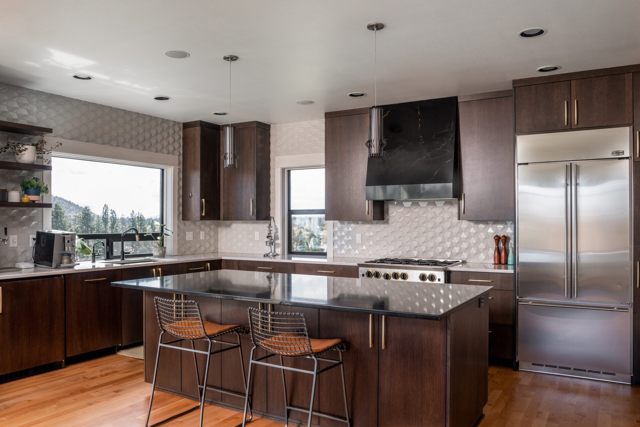 Modern kitchen with dark wood cabinets, a large island, stainless steel refrigerator, and two bar stools, with windows and ceiling lights.