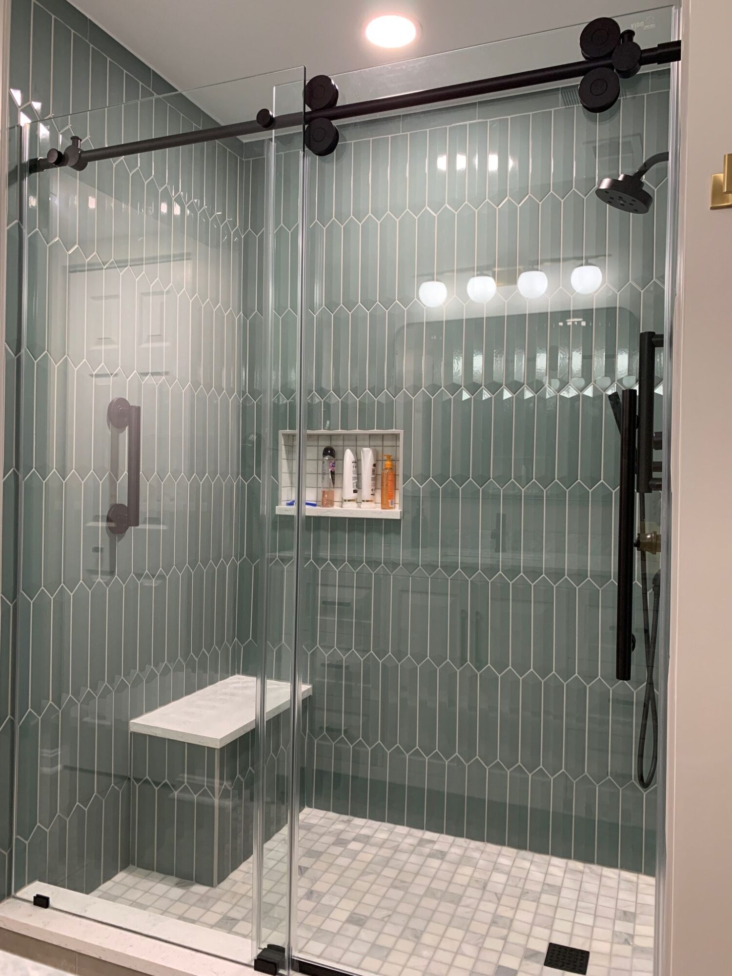 Shower area with glass door, tiled walls, and a showerhead, viewed from the side. Small tiled floor area visible.