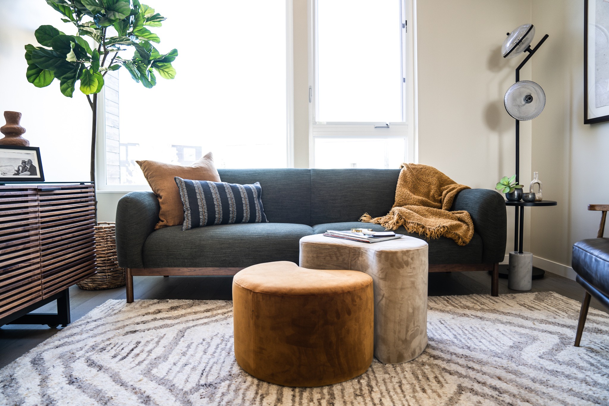Living room with a dark sofa, two round ottomans, a patterned rug, and a large window with sunlight.