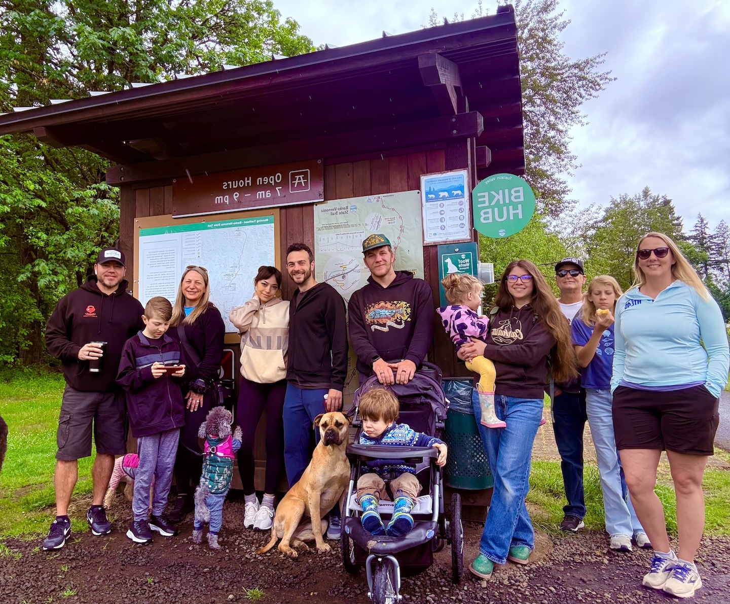 Group of people, including children and adults, standing outdoors near a wooden shelter with informational signs, trees, and cloudy sky in background.