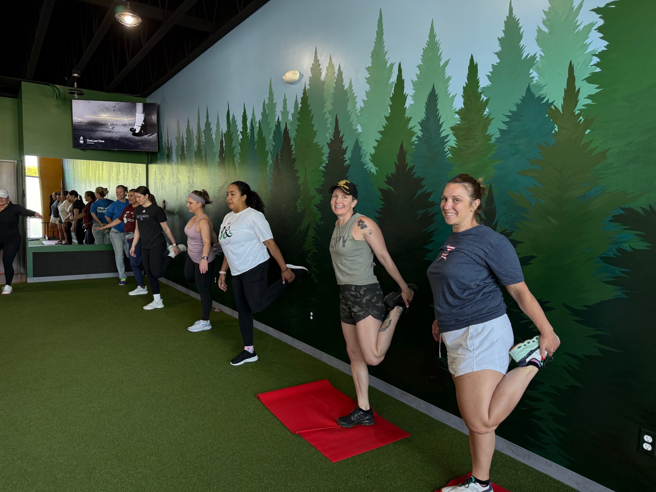 Group of people standing in line indoors against a forest-themed wall, some smiling, with a TV screen in the background.