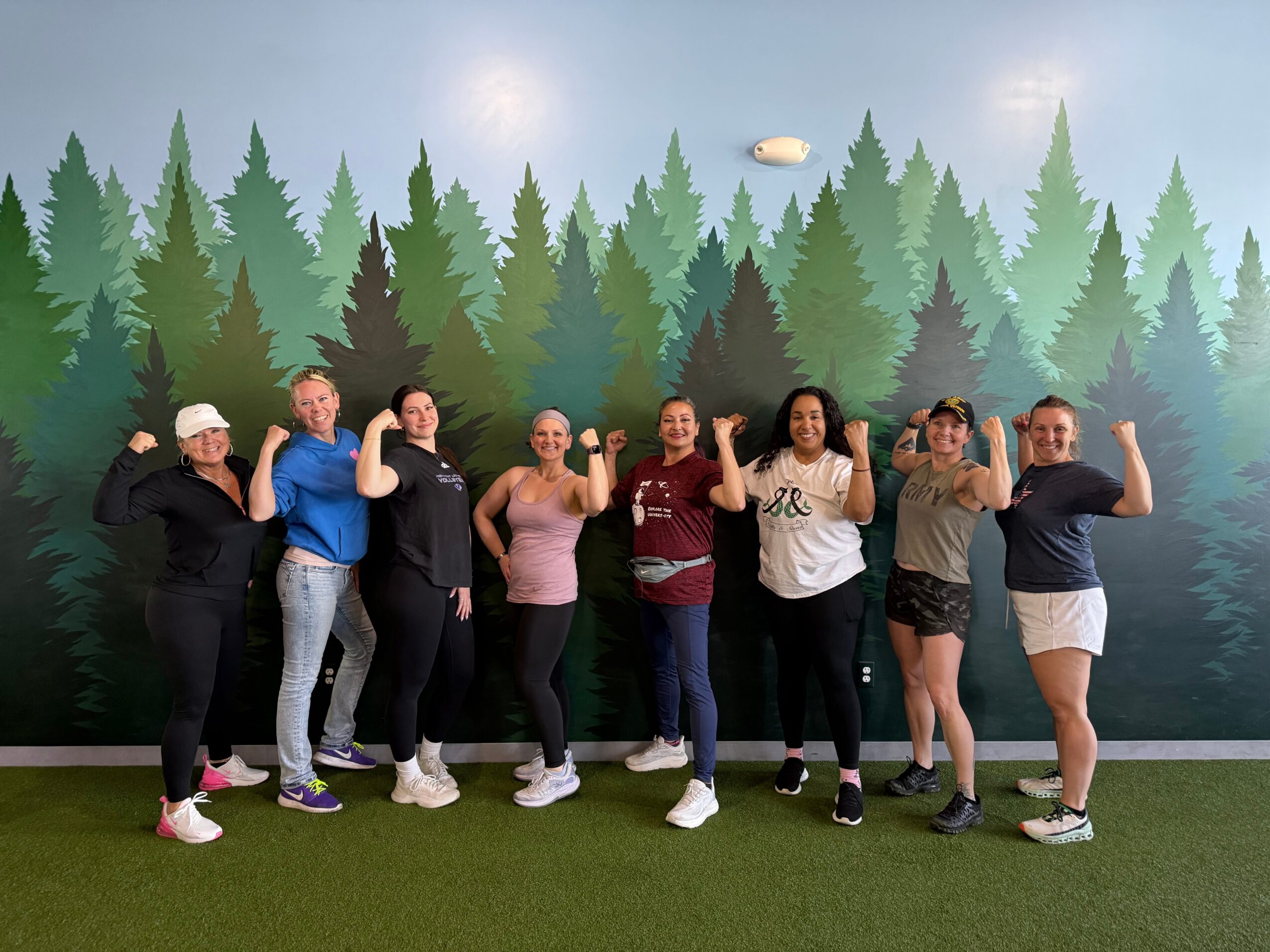 Group of nine people standing outdoors in front of a forest backdrop, posing with arms raised or flexed, smiling.