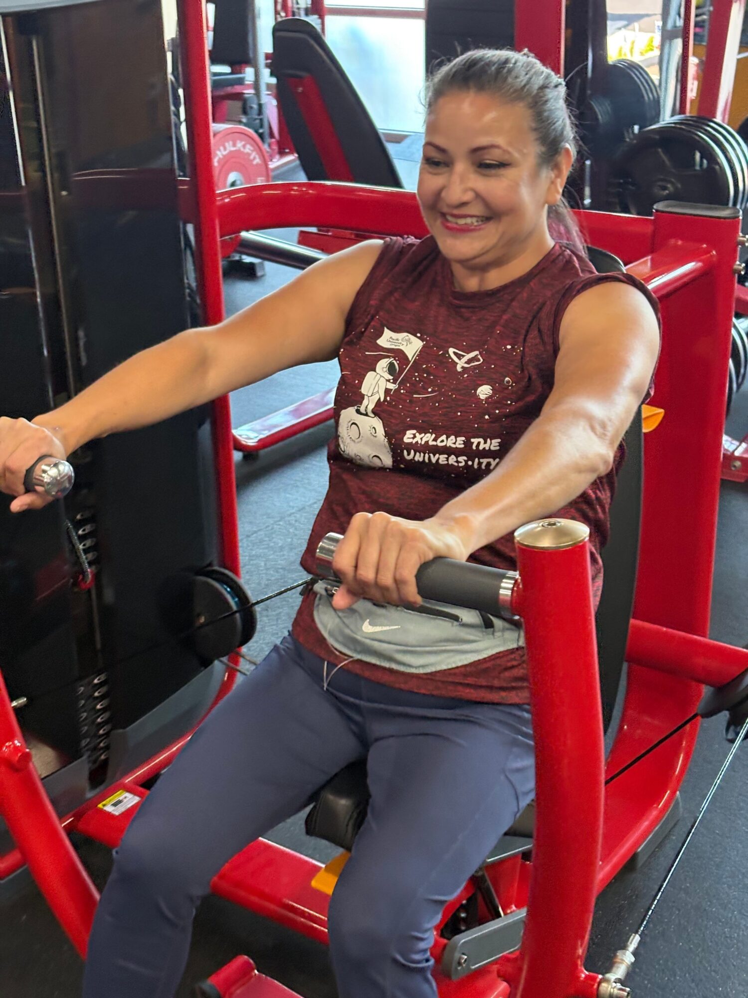 Woman exercising on a red gym machine, holding handles and smiling, in a fitness center.