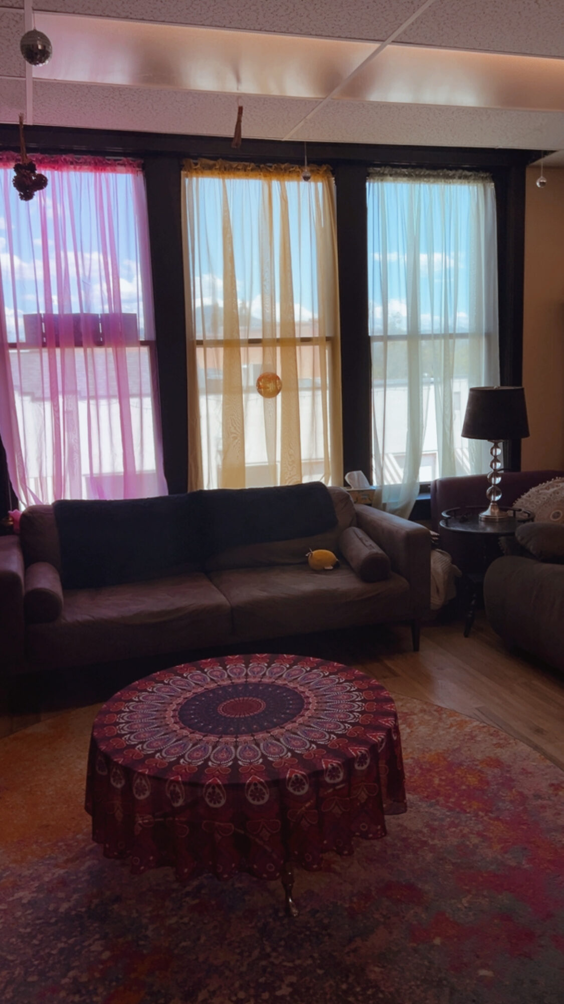 Living room with three colorful curtains, a dark sofa, a round patterned table, and a lamp on a side table.