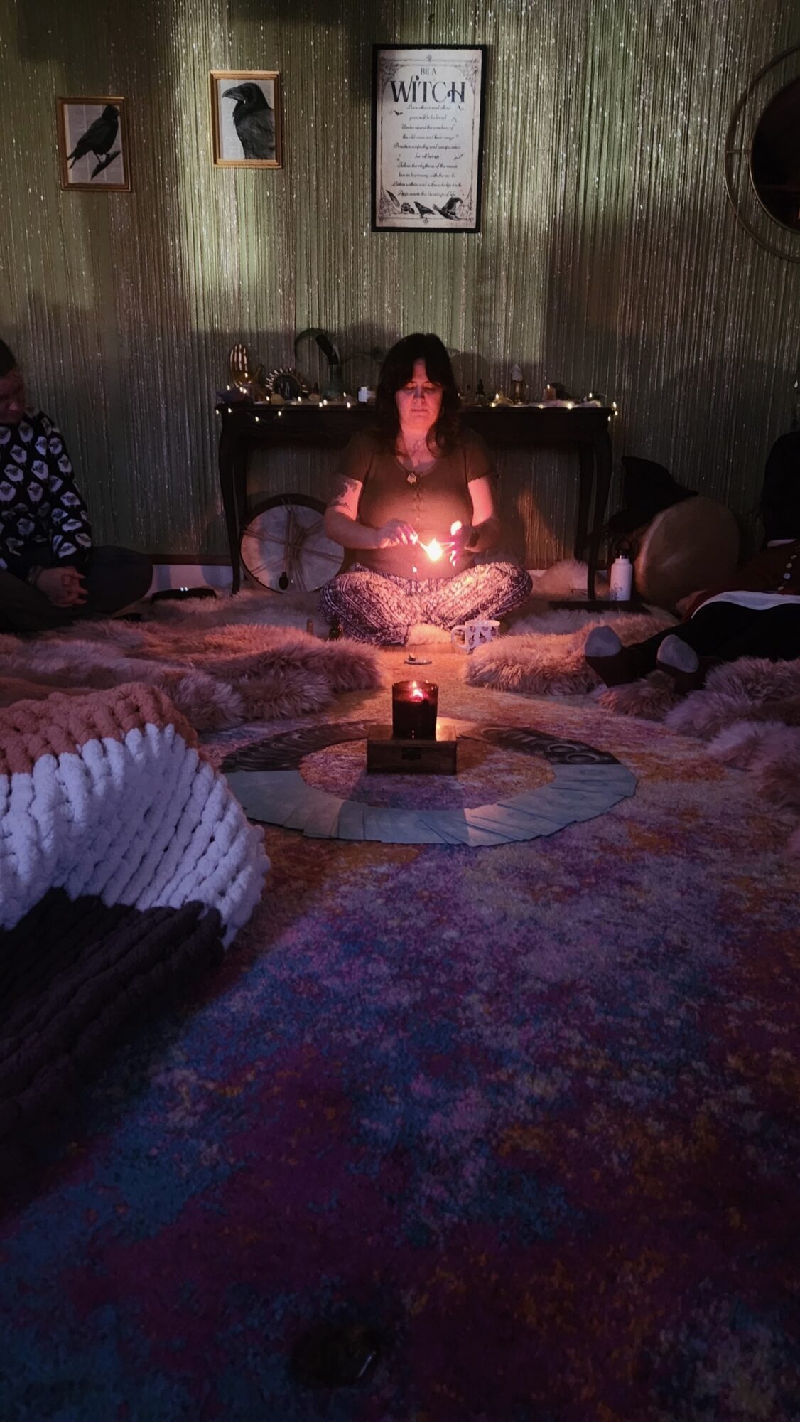Person sitting cross-legged on the floor with a lit candle in front, surrounded by cushions in a dimly lit room.