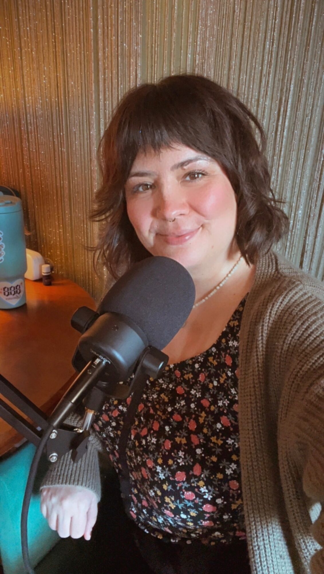 Woman with short brown hair smiling, wearing a floral top and brown cardigan, in front of a microphone.