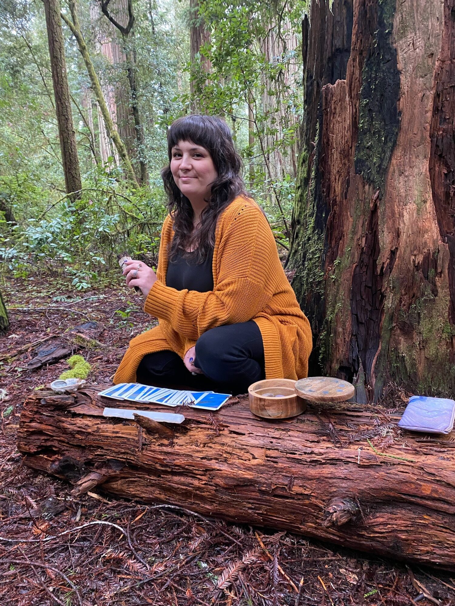 Person with dark hair and a yellow sweater sitting on a fallen log in a forest, with trees and greenery around.