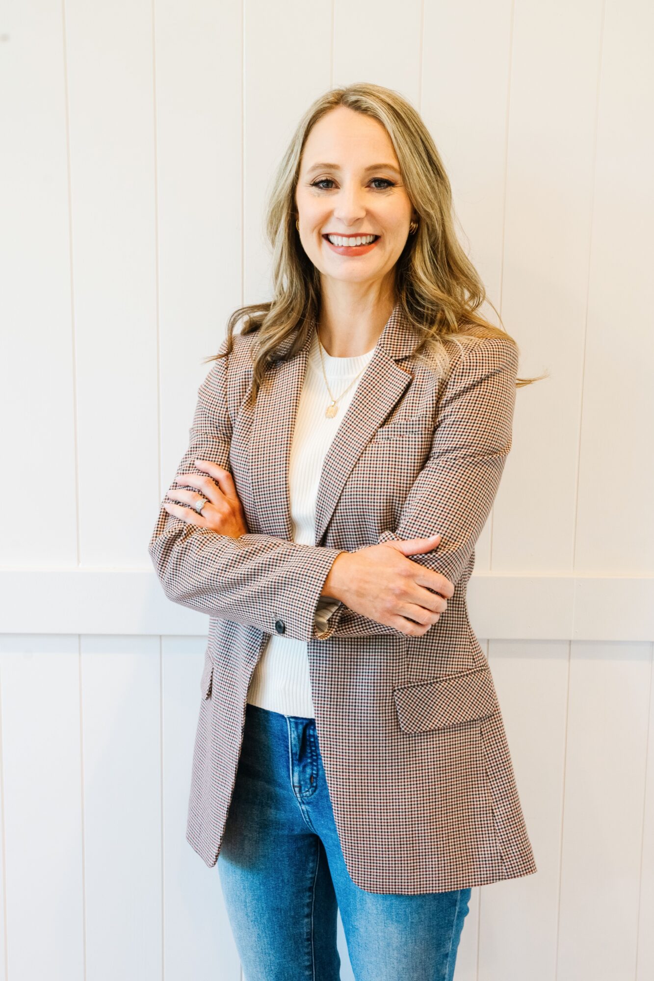 Woman with long wavy hair smiling, arms crossed, wearing a beige blazer and jeans, standing against a white wall.