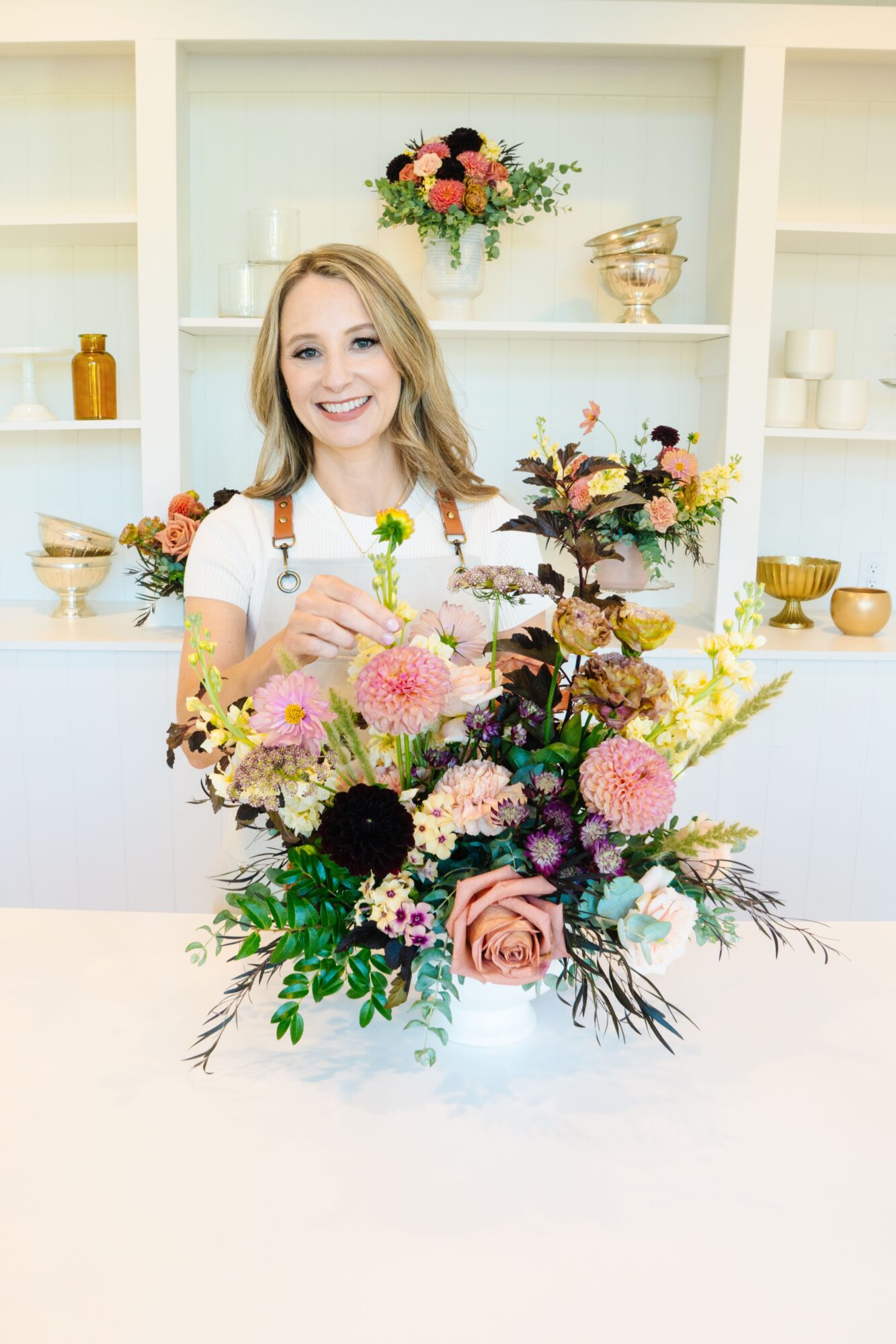 Smiling woman with a stethoscope behind a colorful flower arrangement in a bright room.