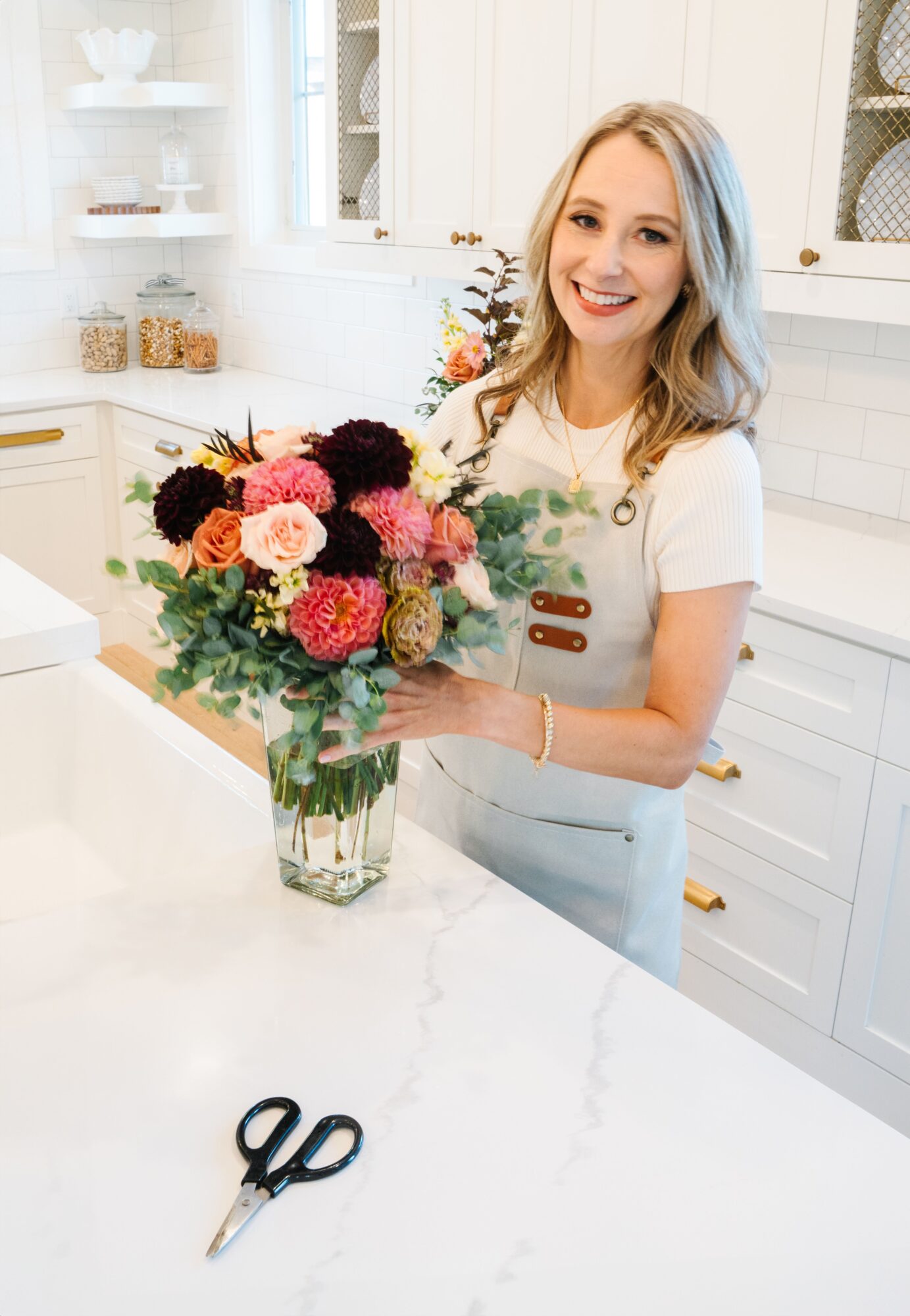Smiling woman with blonde hair holding a colorful flower bouquet in a bright kitchen, with scissors on the counter.