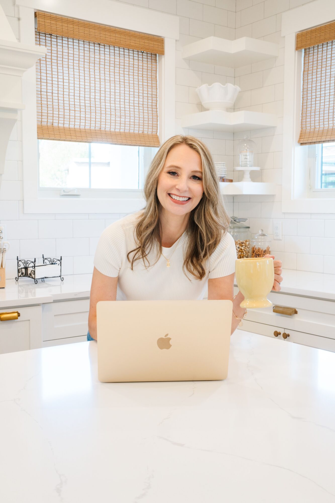 Smiling woman sitting at a kitchen counter with a laptop, white walls, and windows with blinds behind her.