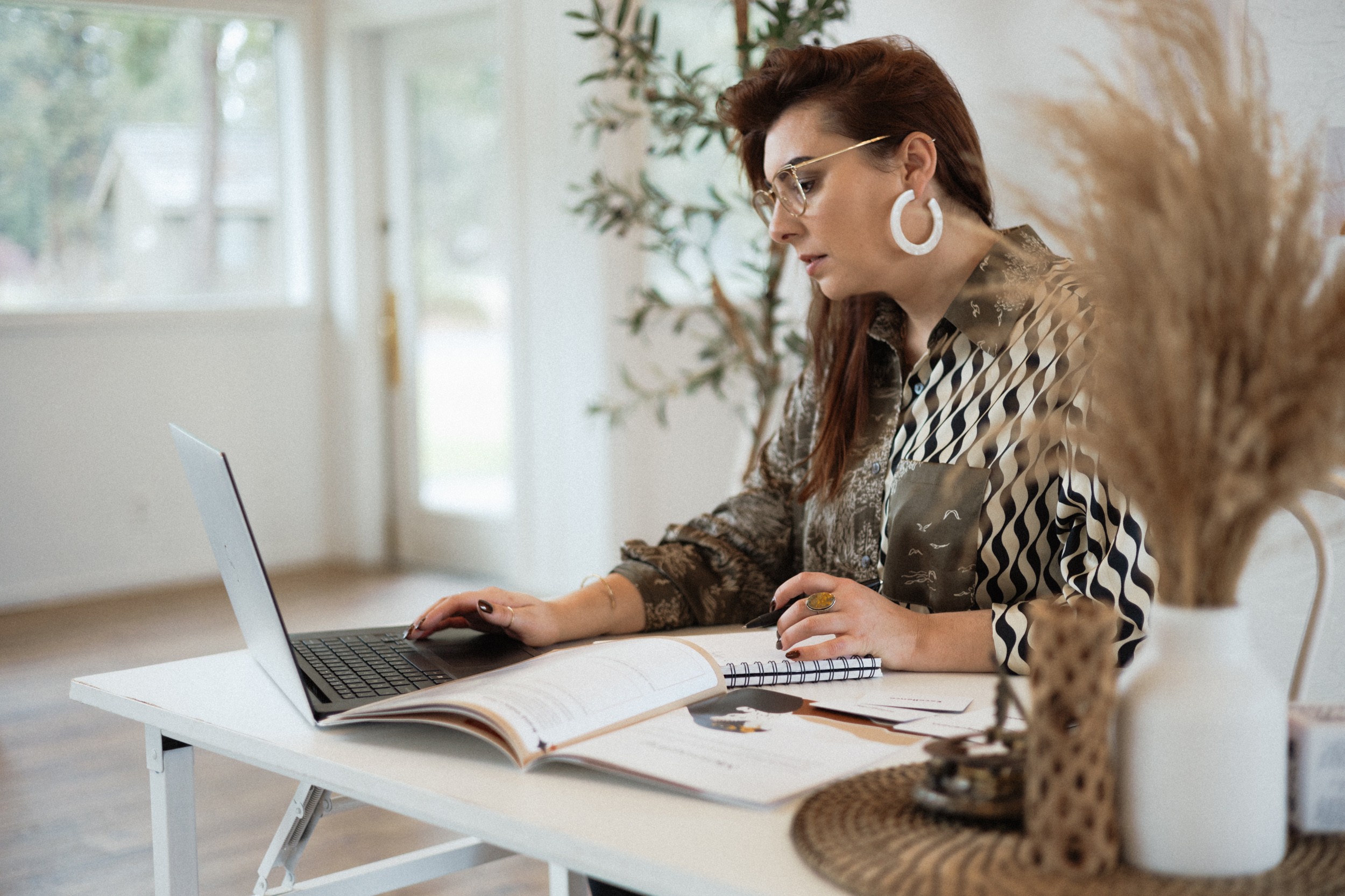 Woman with glasses and earrings working on a laptop at a white desk, with open books and plants nearby.