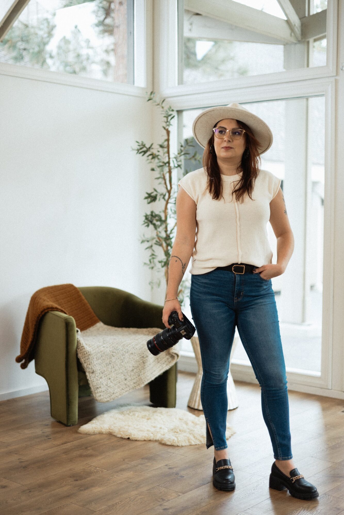 Woman in a white top, jeans, and wide-brimmed hat stands indoors holding a camera, with a green armchair and window behind.