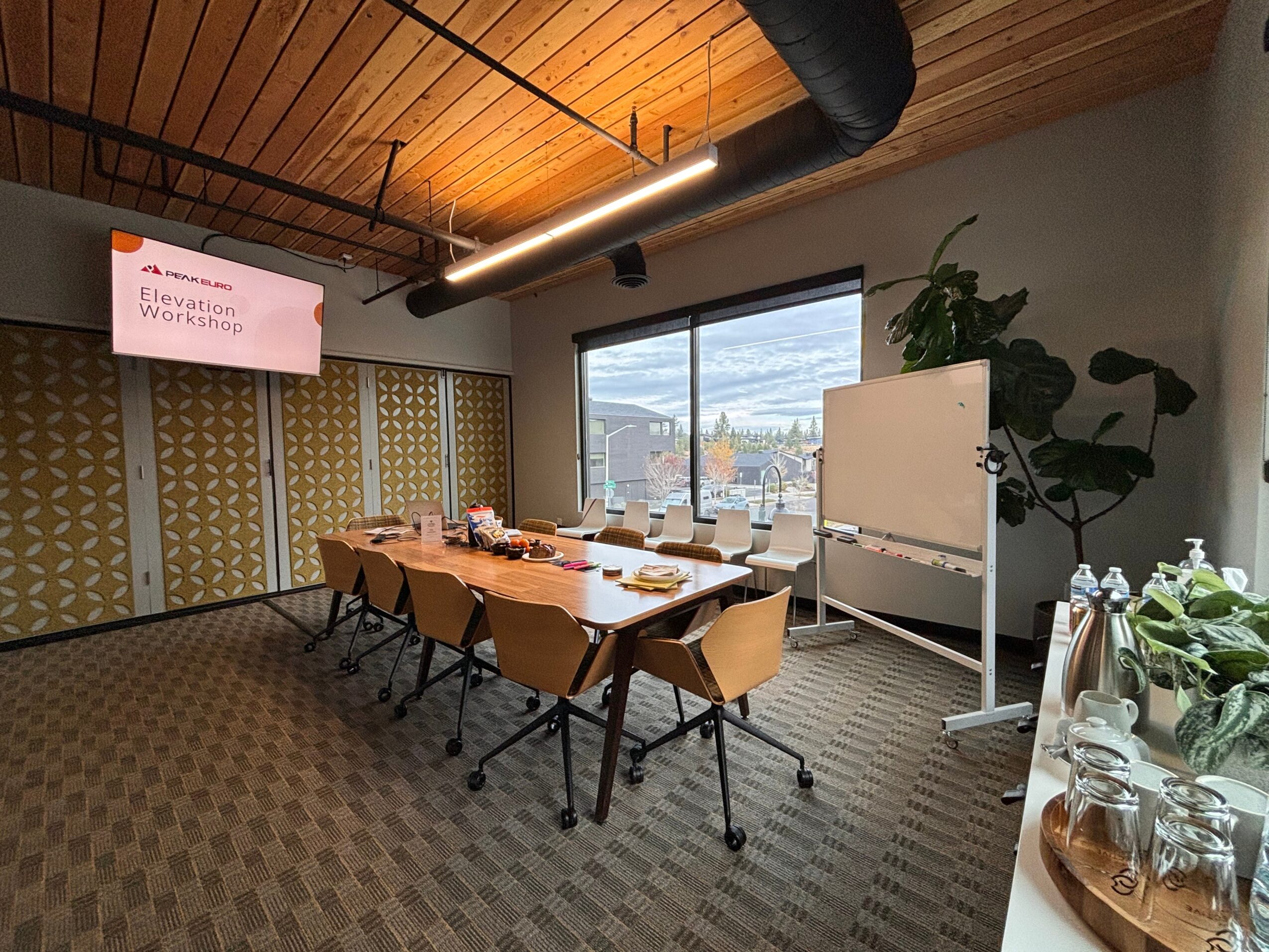 Meeting room with a long table, chairs, window, whiteboard, and plants, under a wooden ceiling with a projector screen.
