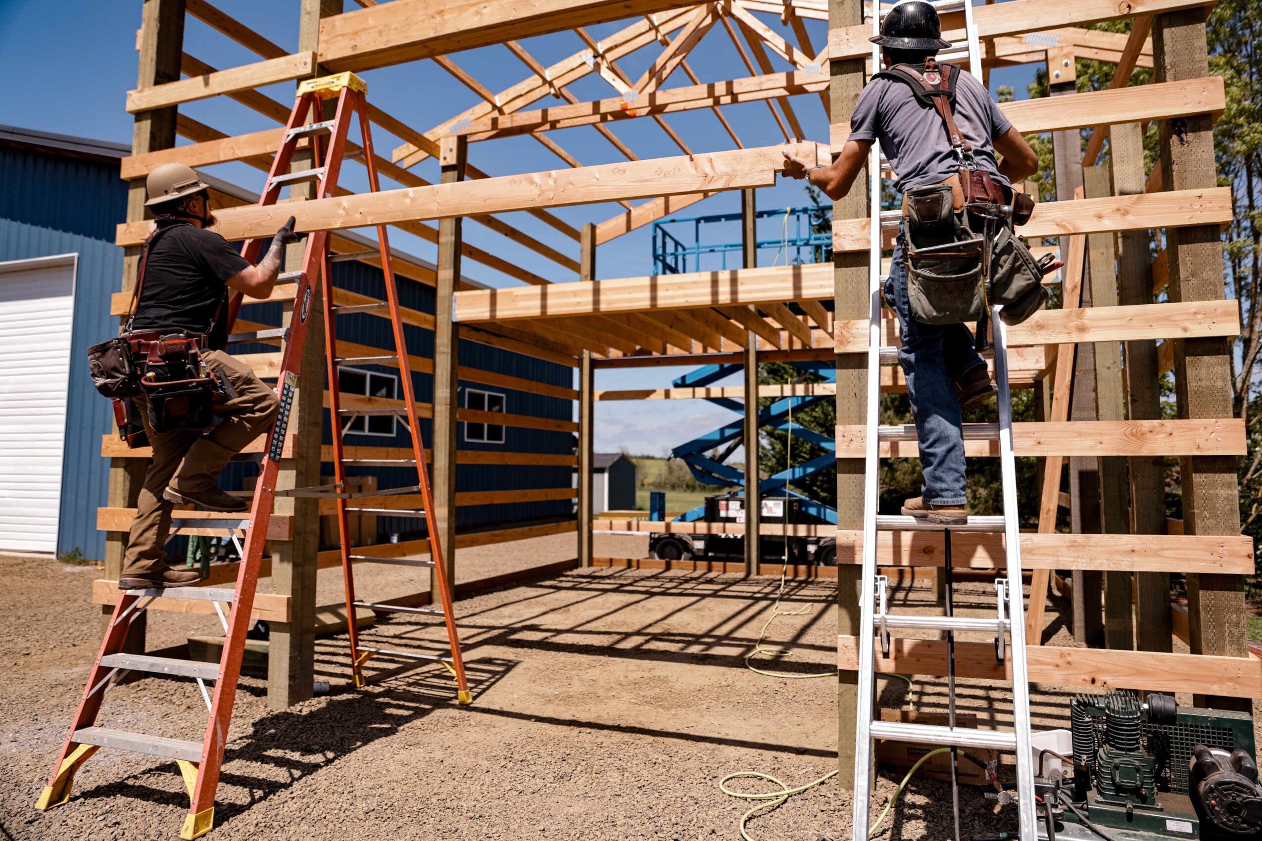 Two construction workers building a wooden frame structure outdoors, one on a ladder and one on a scaffold, with tools nearby.