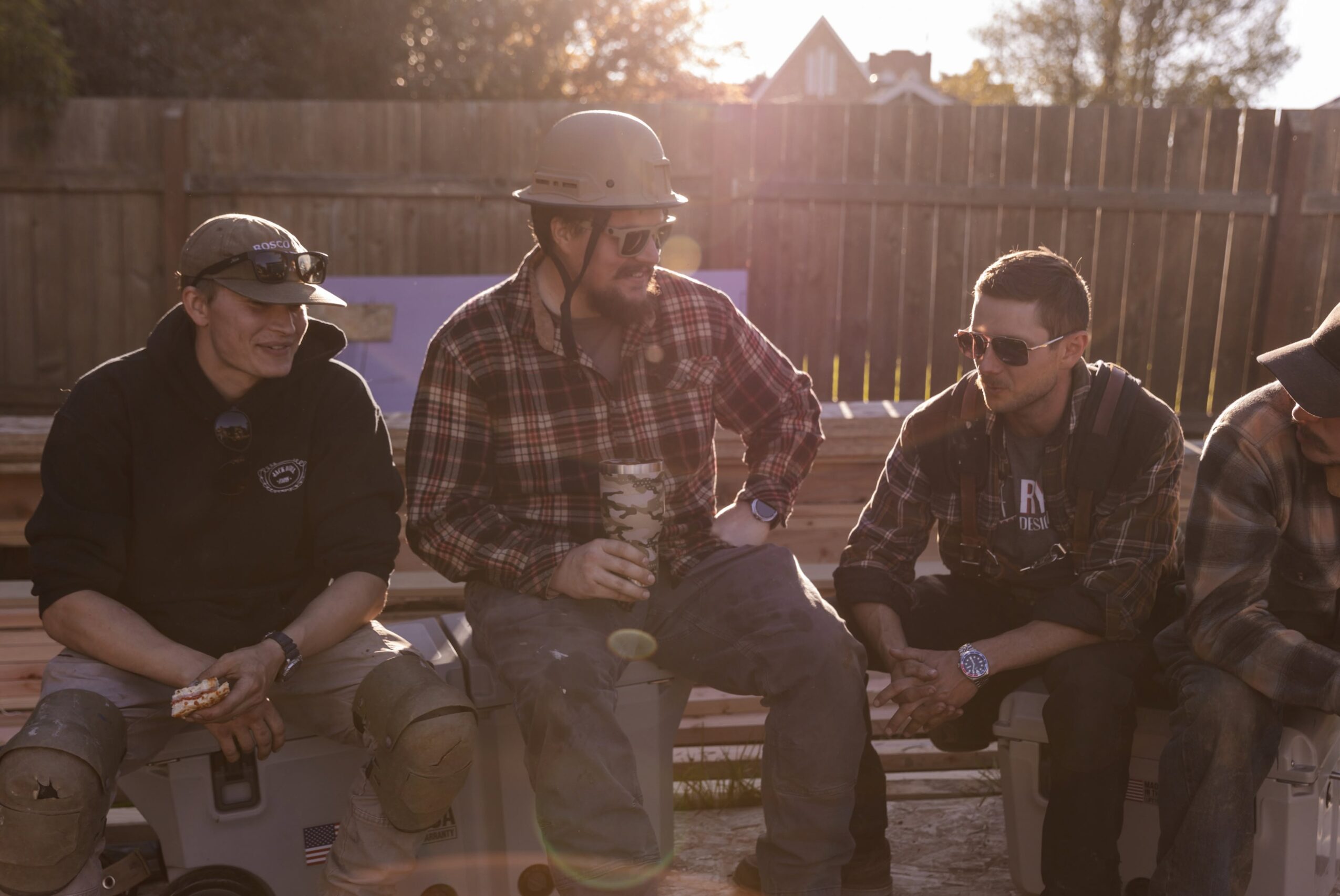 Four people sitting on a bench outdoors, one wearing a helmet, all wearing sunglasses, with a wooden fence in the background.