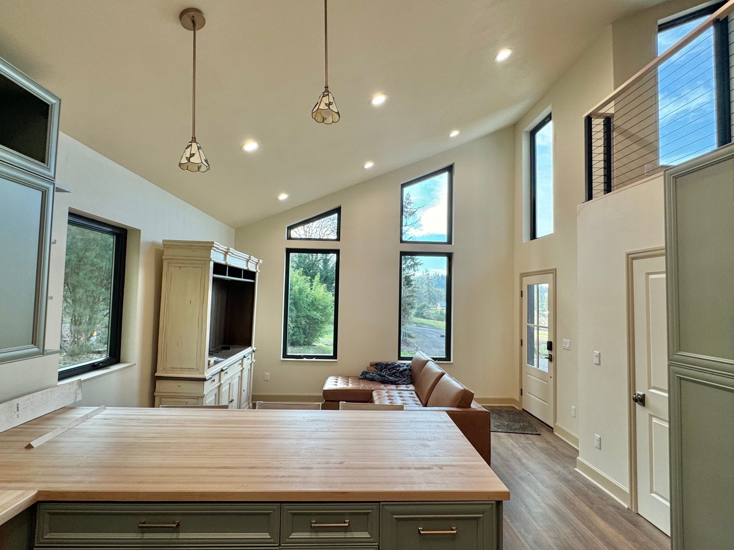 Living room with large windows, a brown sofa, and a wooden table, illuminated by pendant lights and natural light.