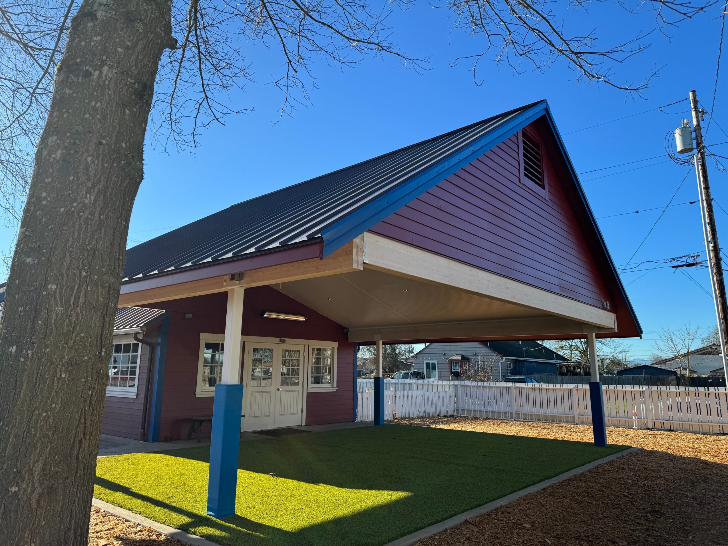 Small building with a sloped metal roof, purple walls, and a covered porch area, surrounded by grass and a white fence.