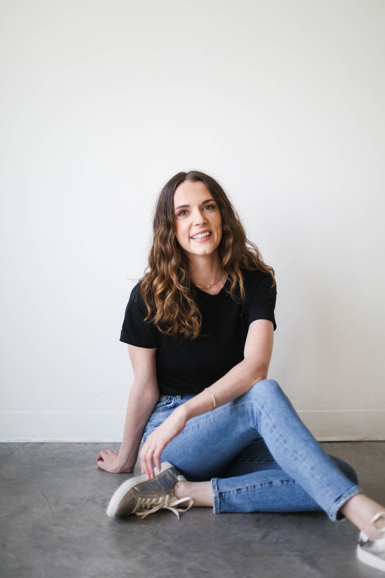 Young woman with wavy hair sitting on the floor, smiling, wearing a black t-shirt and blue jeans, against a plain white wall.