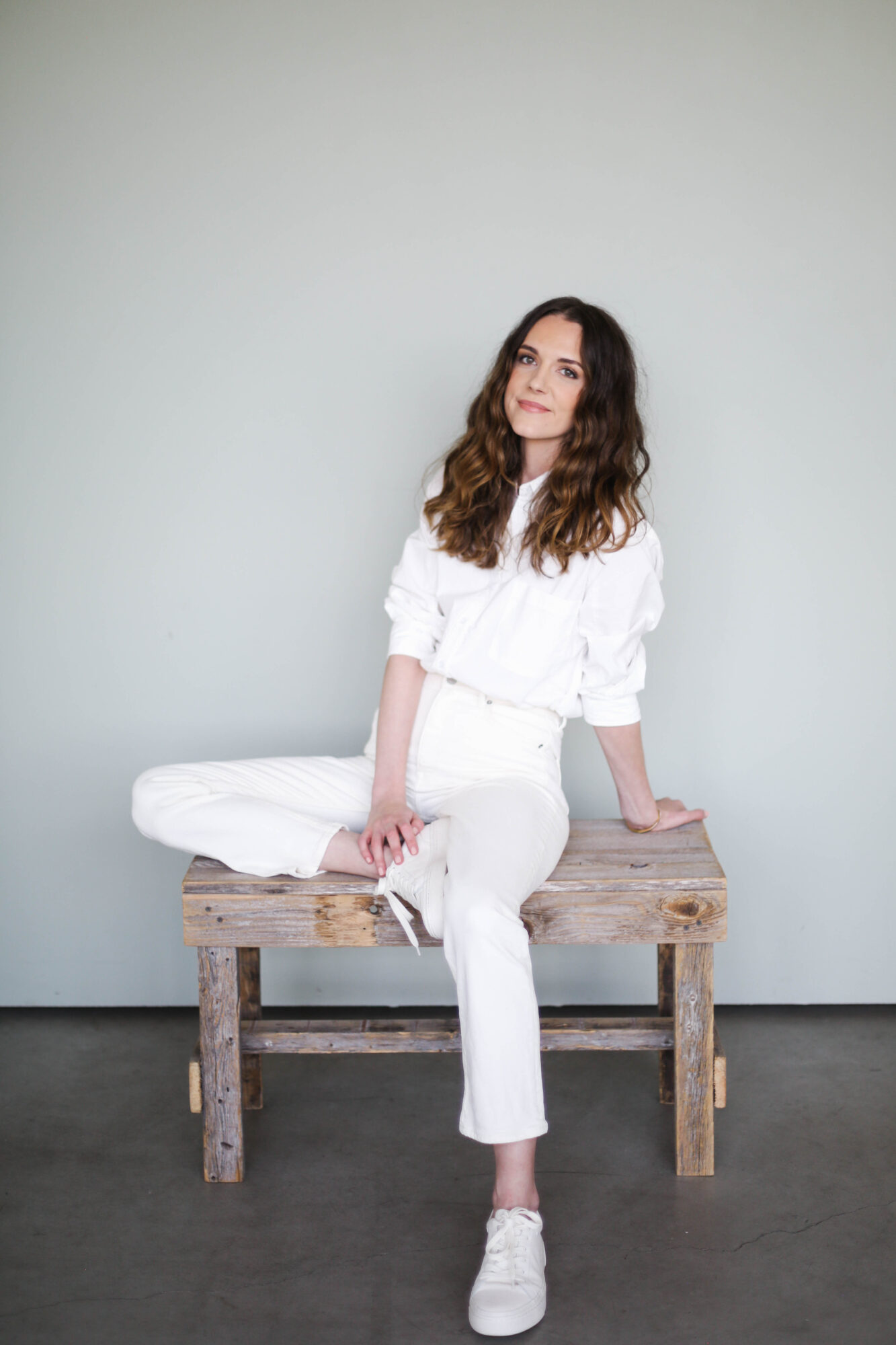 Woman with wavy hair sitting on a wooden bench against a plain light-colored wall, wearing white clothing and sneakers.