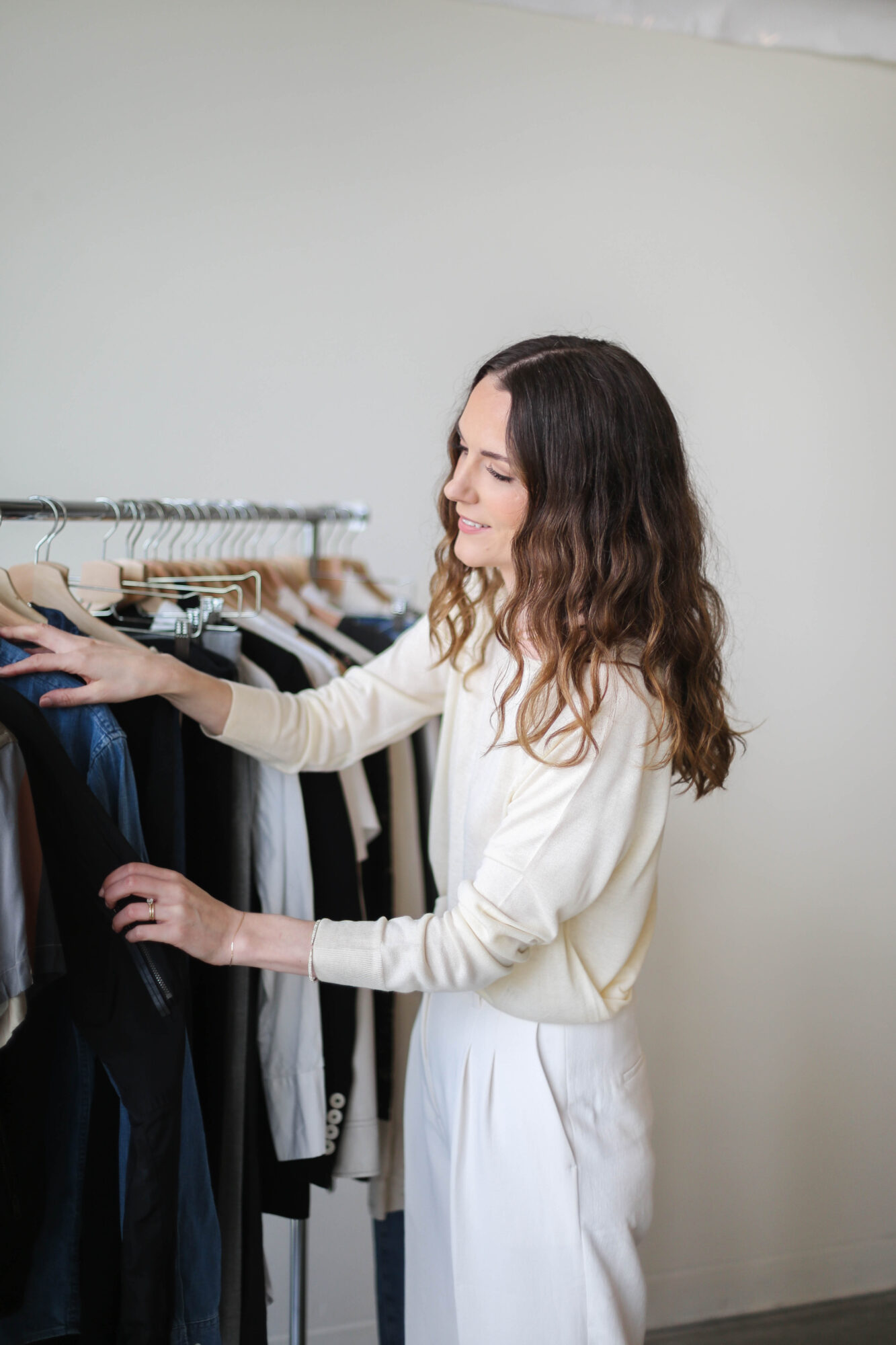 Woman with long wavy hair browsing clothes on a rack in a store, wearing a white outfit, neutral background.
