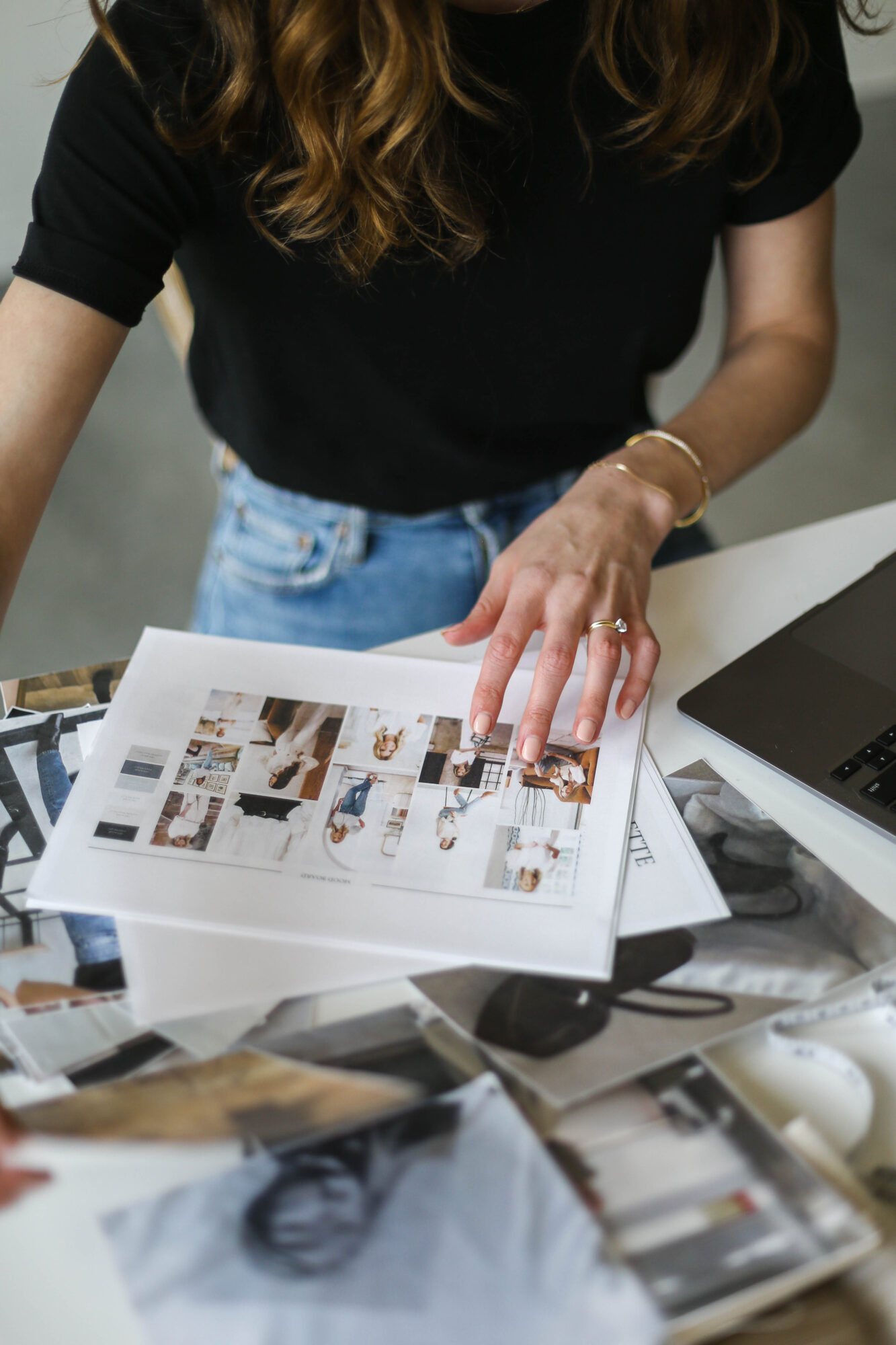 Person with long hair wearing a black shirt and jeans, looking at printed images on a desk, with a laptop nearby.