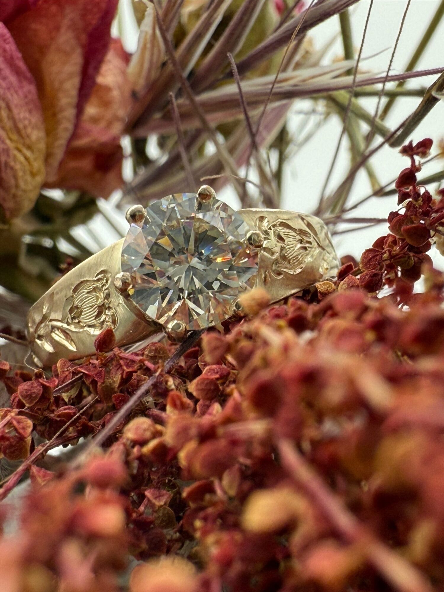 A diamond ring with a large central gemstone surrounded by smaller stones, placed among dried flowers and foliage.