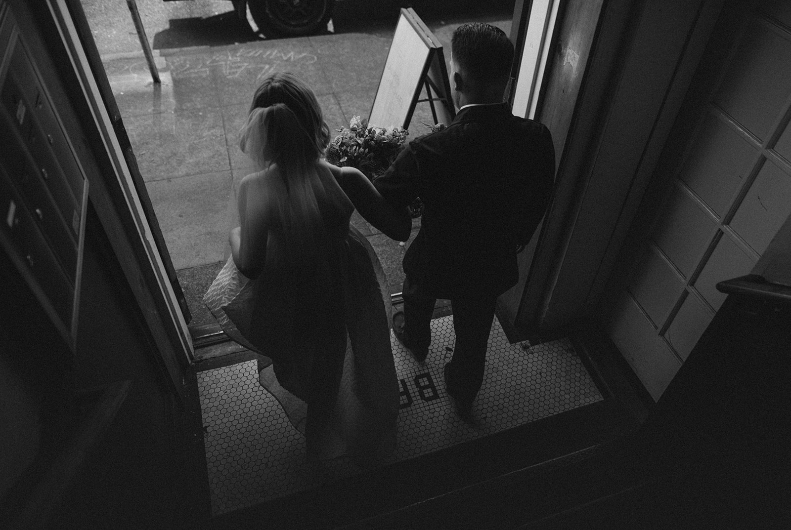 Two people, a woman and a man, walk out of a building onto a street, holding hands, seen from above.