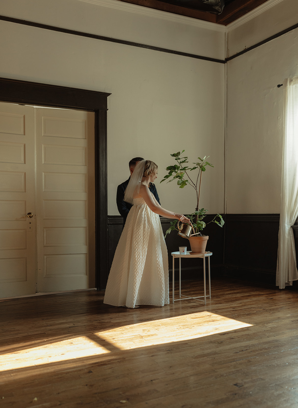 A bride and groom stand together in a room with wooden floors, a potted plant, and a window with curtains.