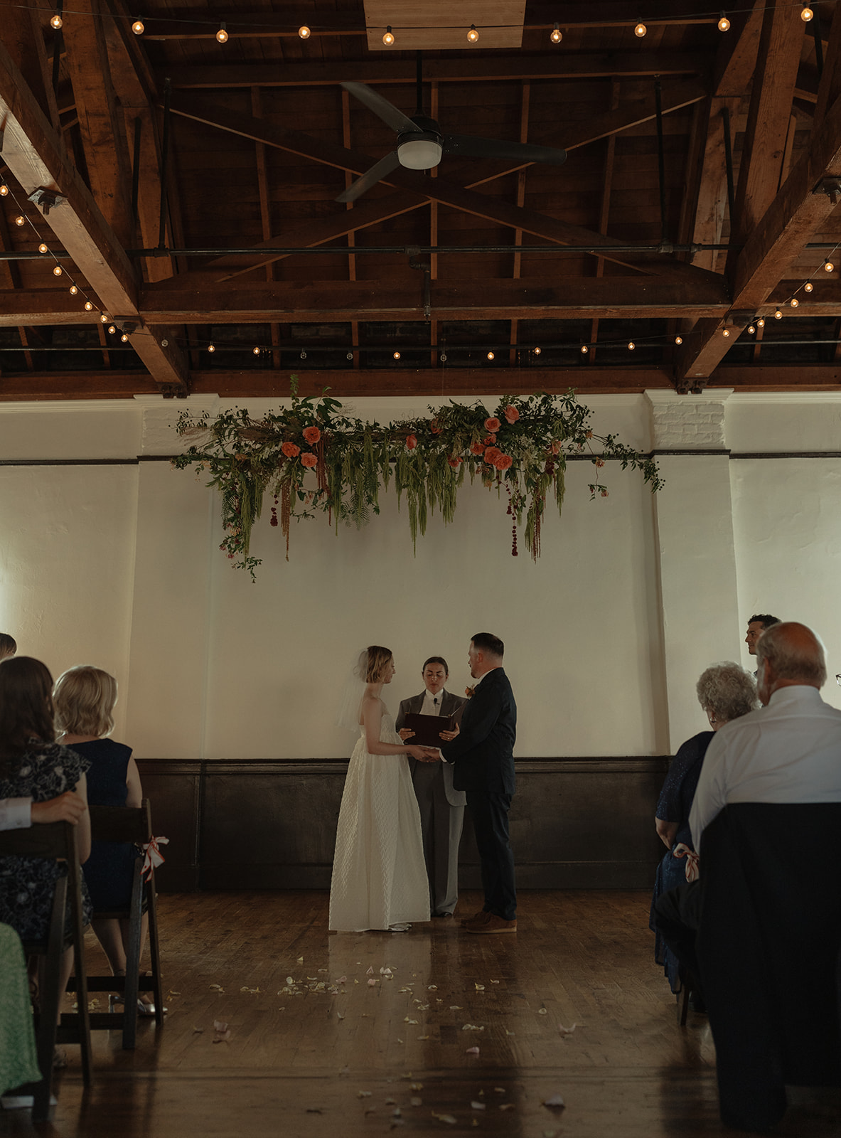 A wedding ceremony with a bride and groom standing before an officiant, under a floral arrangement, in a spacious hall.