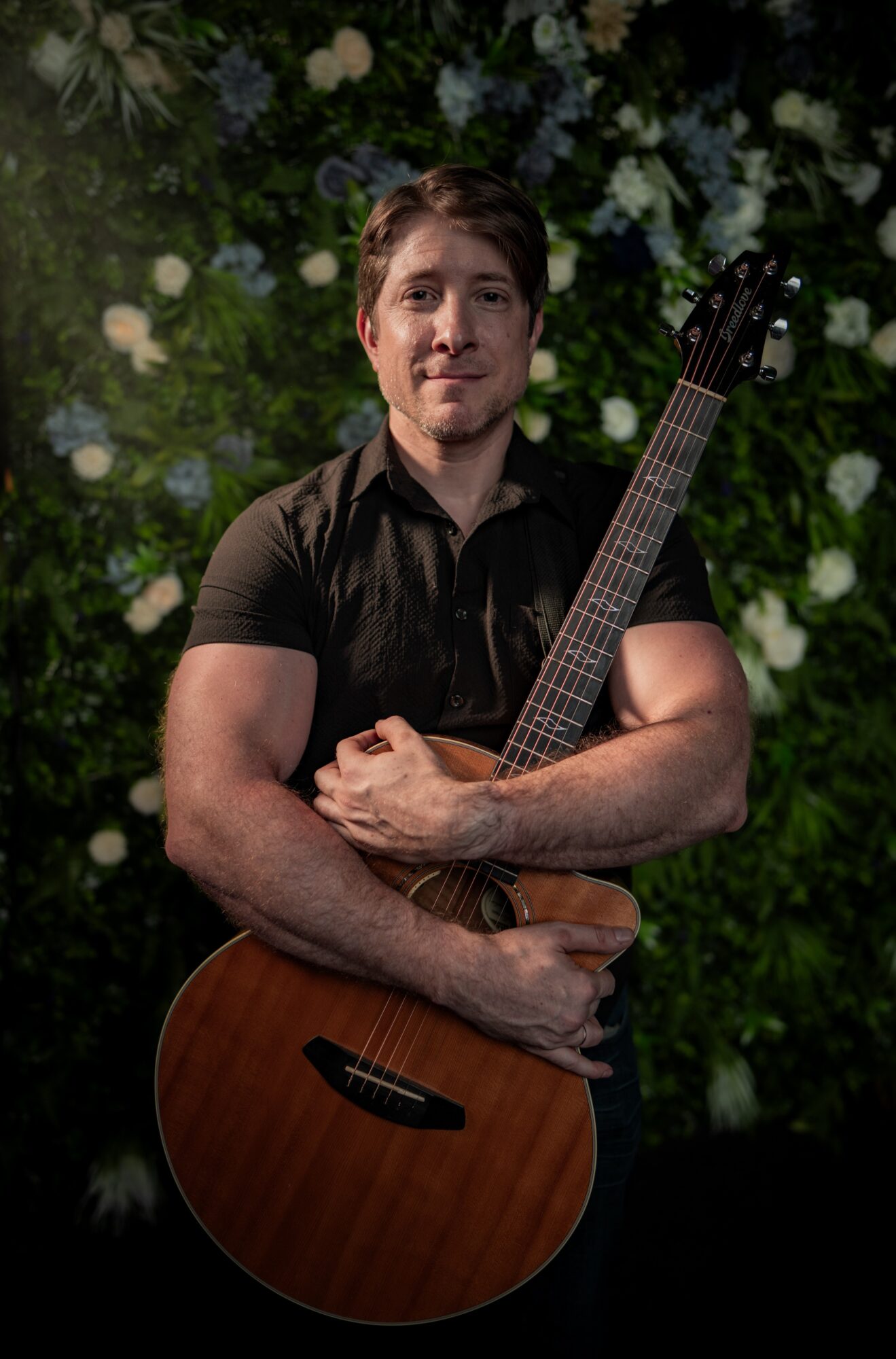 Man holding an acoustic guitar in front of a leafy background, smiling, with arms crossed over the guitar.