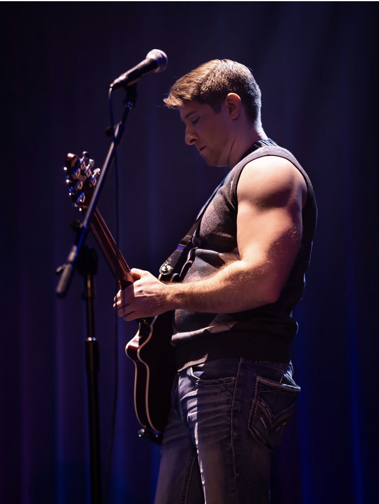 Young man playing guitar on stage with a microphone, dark background, focused expression, wearing sleeveless shirt and jeans.