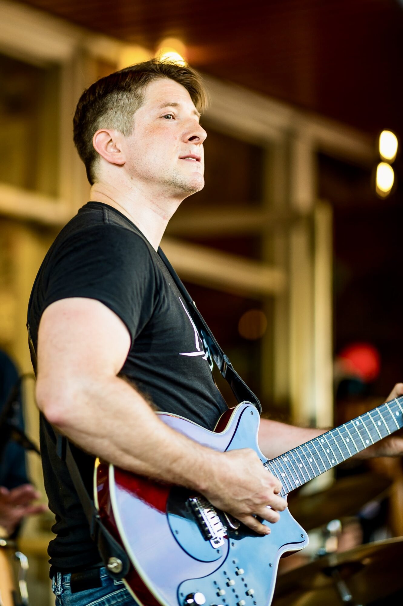 Young man playing an electric guitar indoors, wearing a black t-shirt, with a focused expression, background with warm lighting.