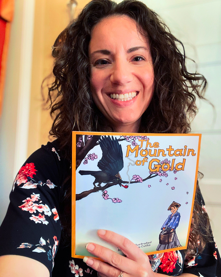 Woman with curly hair smiling holding a book titled 'The Mountain of Gold' in front of a light-colored wall.