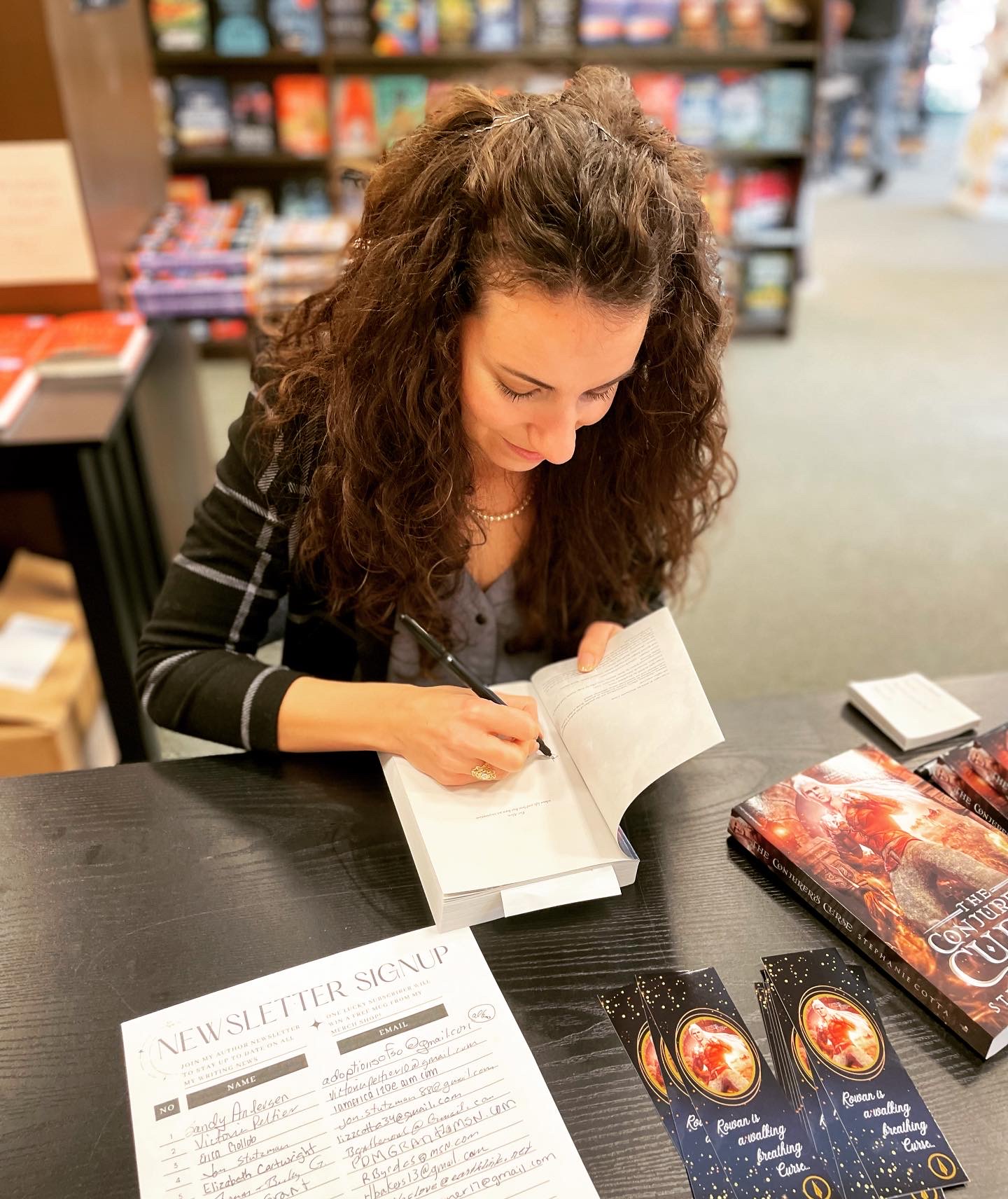 Woman with curly hair reading and writing in a book at a table in a bookstore, with books and magazines nearby.