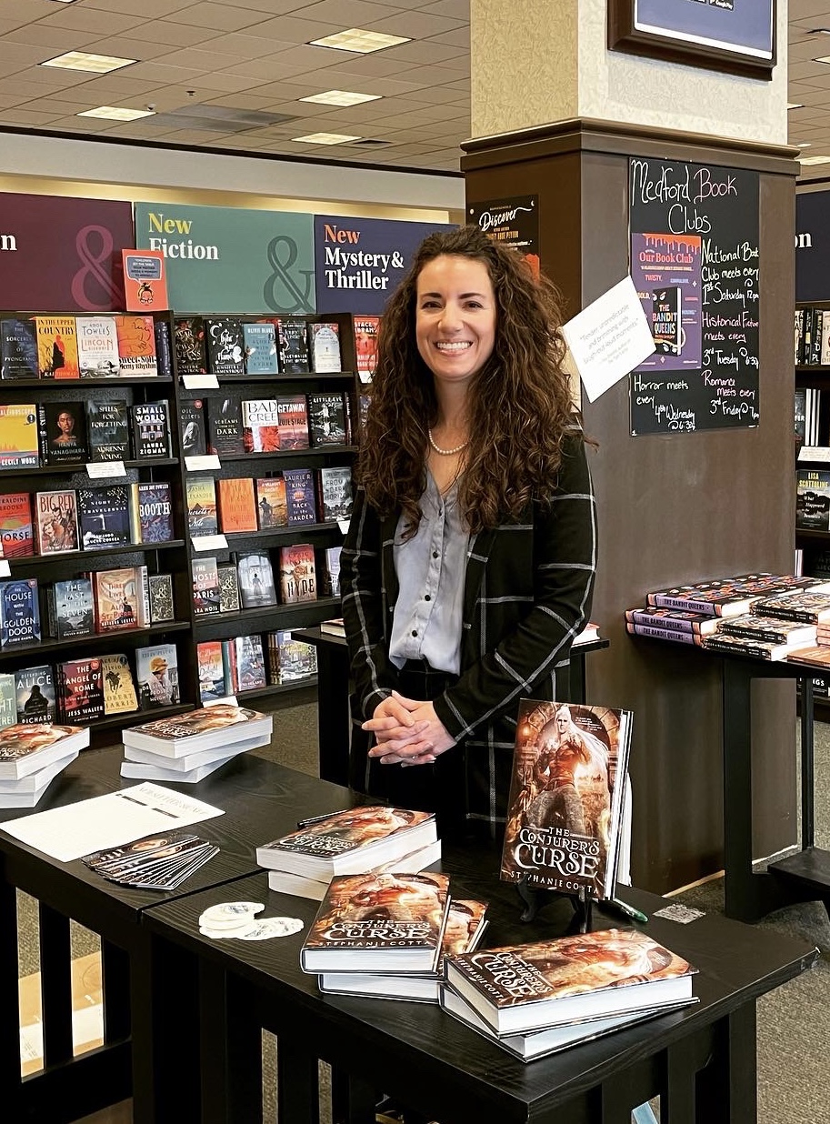 Woman with long curly hair standing behind a table of books in a bookstore, smiling, with shelves of books behind her.