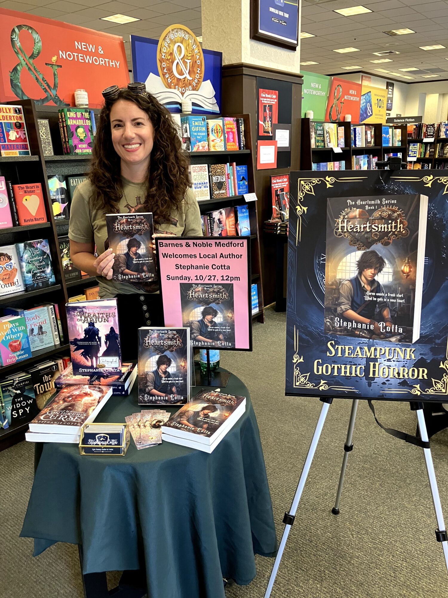Woman with curly hair standing behind a book display table in a bookstore, promoting a book signing event. Books are arranged on the table and shelves behind her.