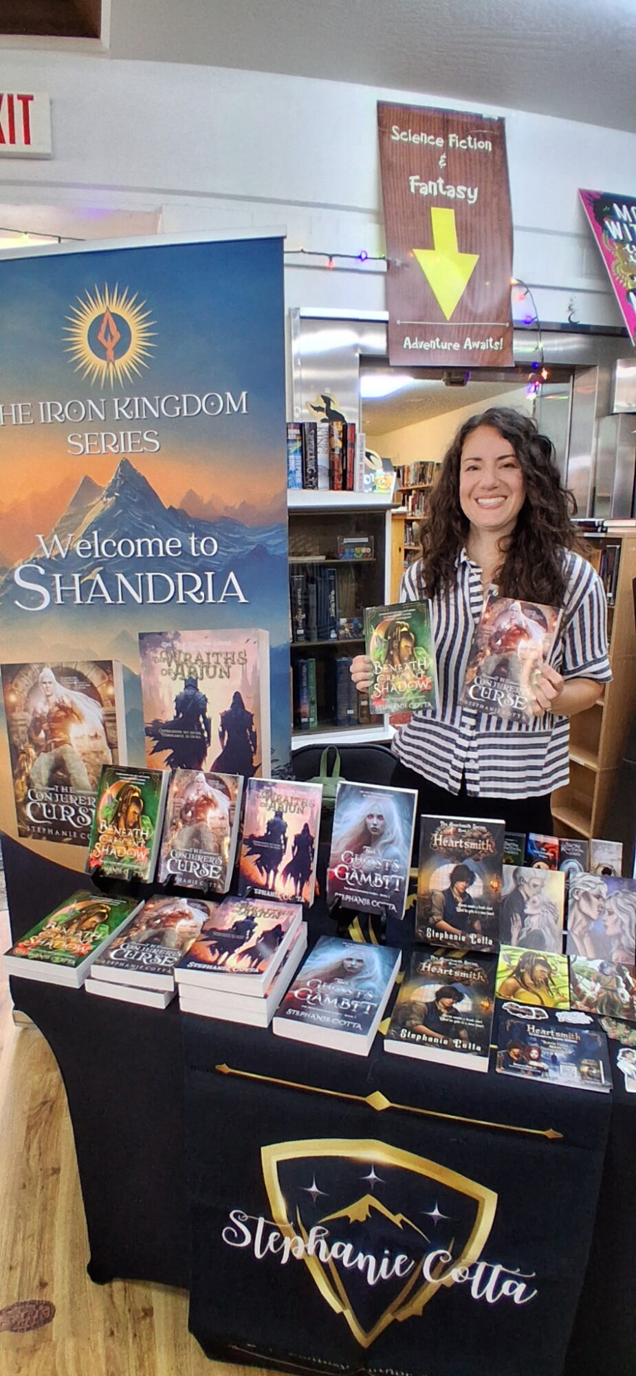 Woman with curly hair standing behind a table of fantasy books at a book signing event, smiling and holding a book.