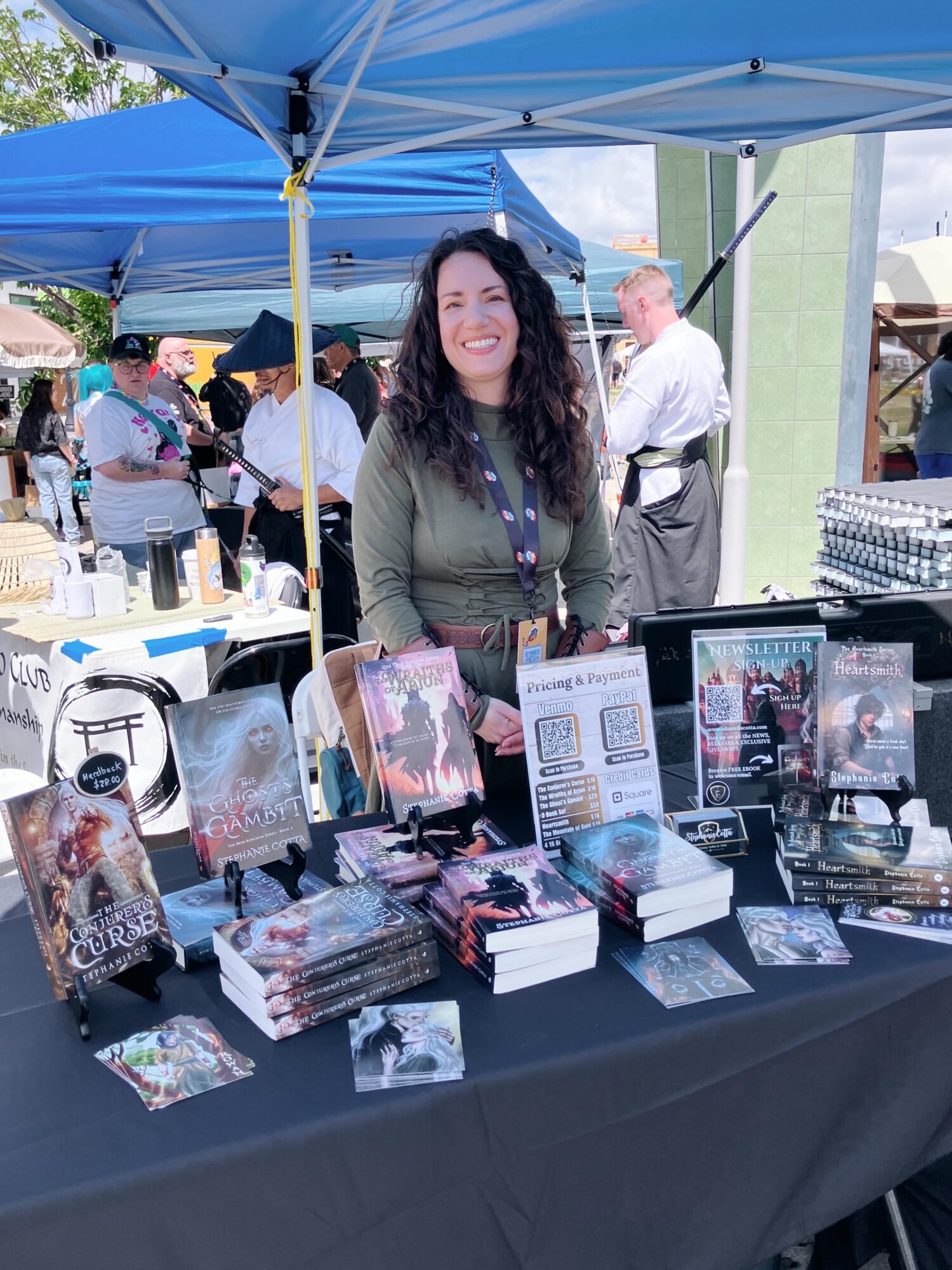 Woman standing behind a table with books and promotional materials at an outdoor event, tents and people in background.