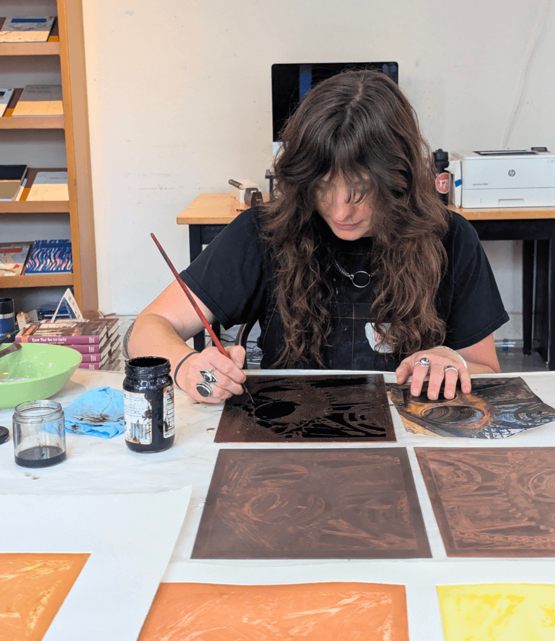 Person with long curly hair painting on a black surface at a table with art supplies, books, and printed artwork.