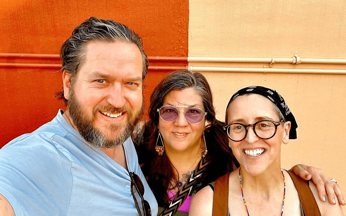 Three people smiling outdoors, one man and two women, standing close together, with a colorful background.