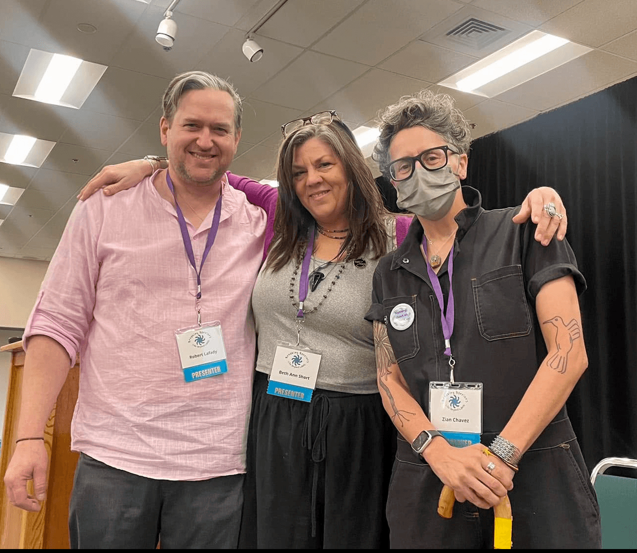 Three people standing together indoors, smiling, with conference badges and lanyards, one wearing a face mask, in a well-lit room.