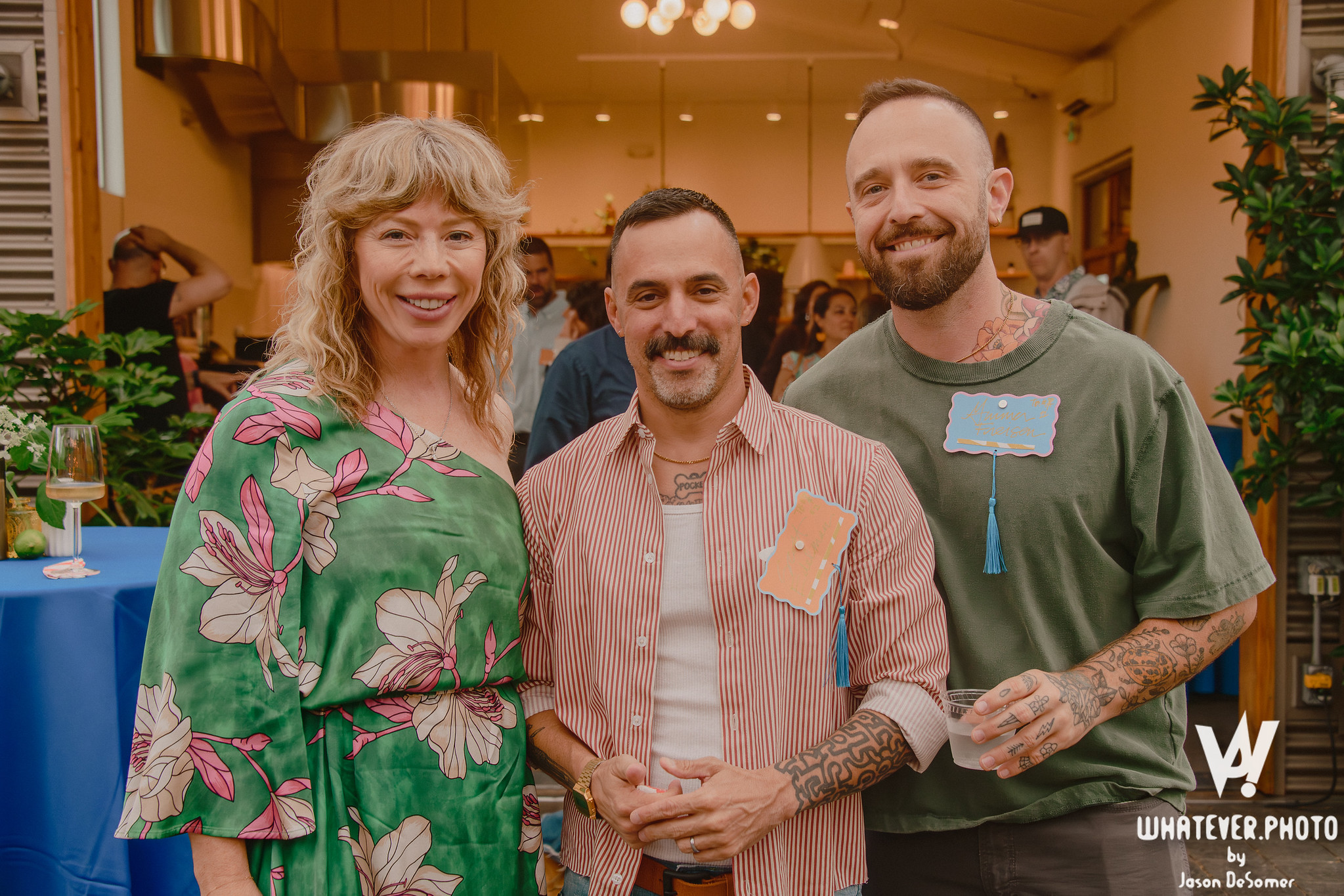 Three people smiling at a social event, standing indoors with a crowd and decorated tables in the background.