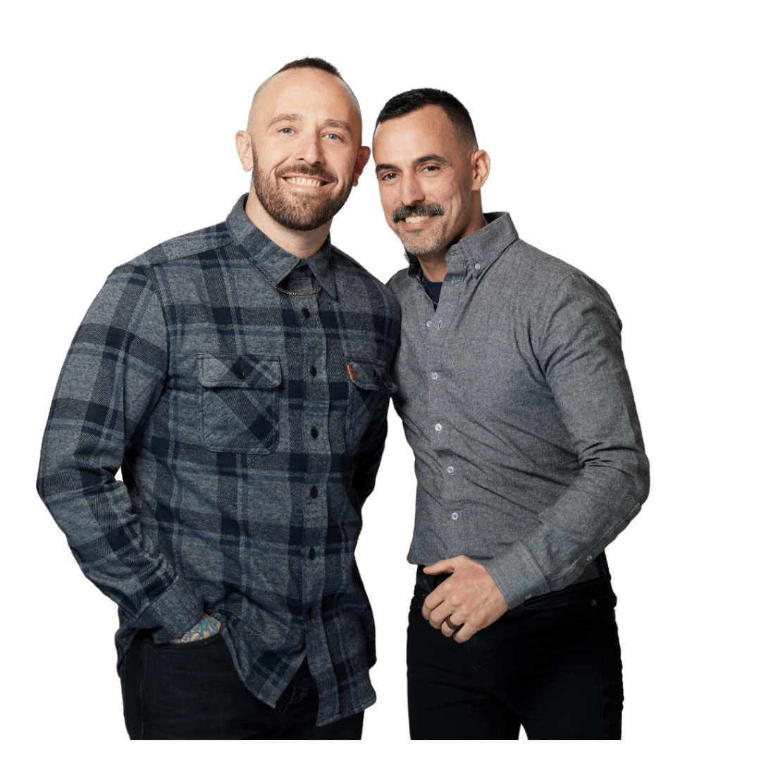 Two men stand close together, smiling, against a black background. One wears a plaid shirt, the other a gray shirt.