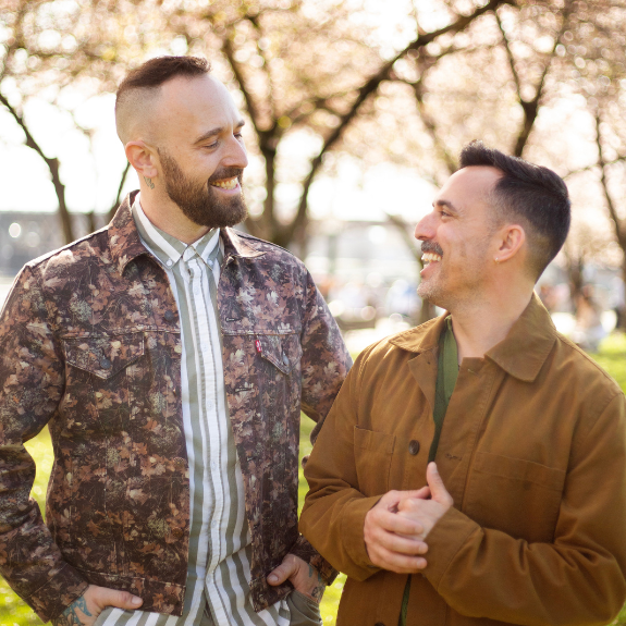 Two men smiling and talking outdoors with trees in the background.