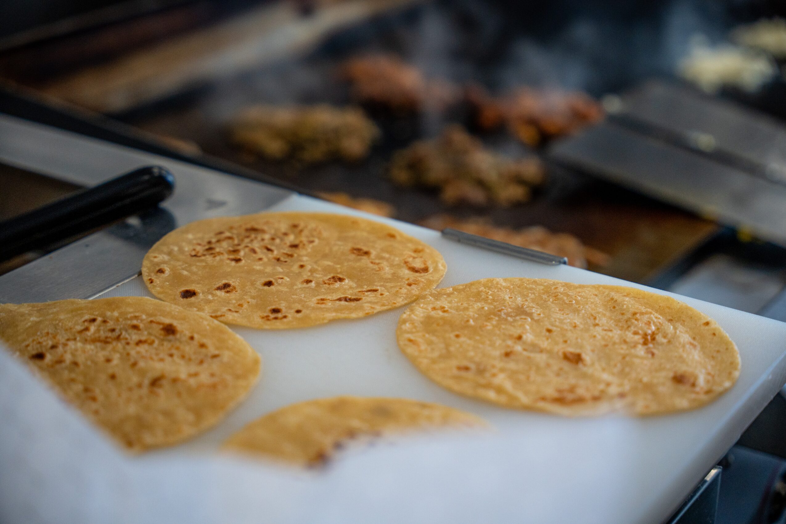 Three flatbreads on a white surface with a grill and food in the background, cooking on a stovetop.