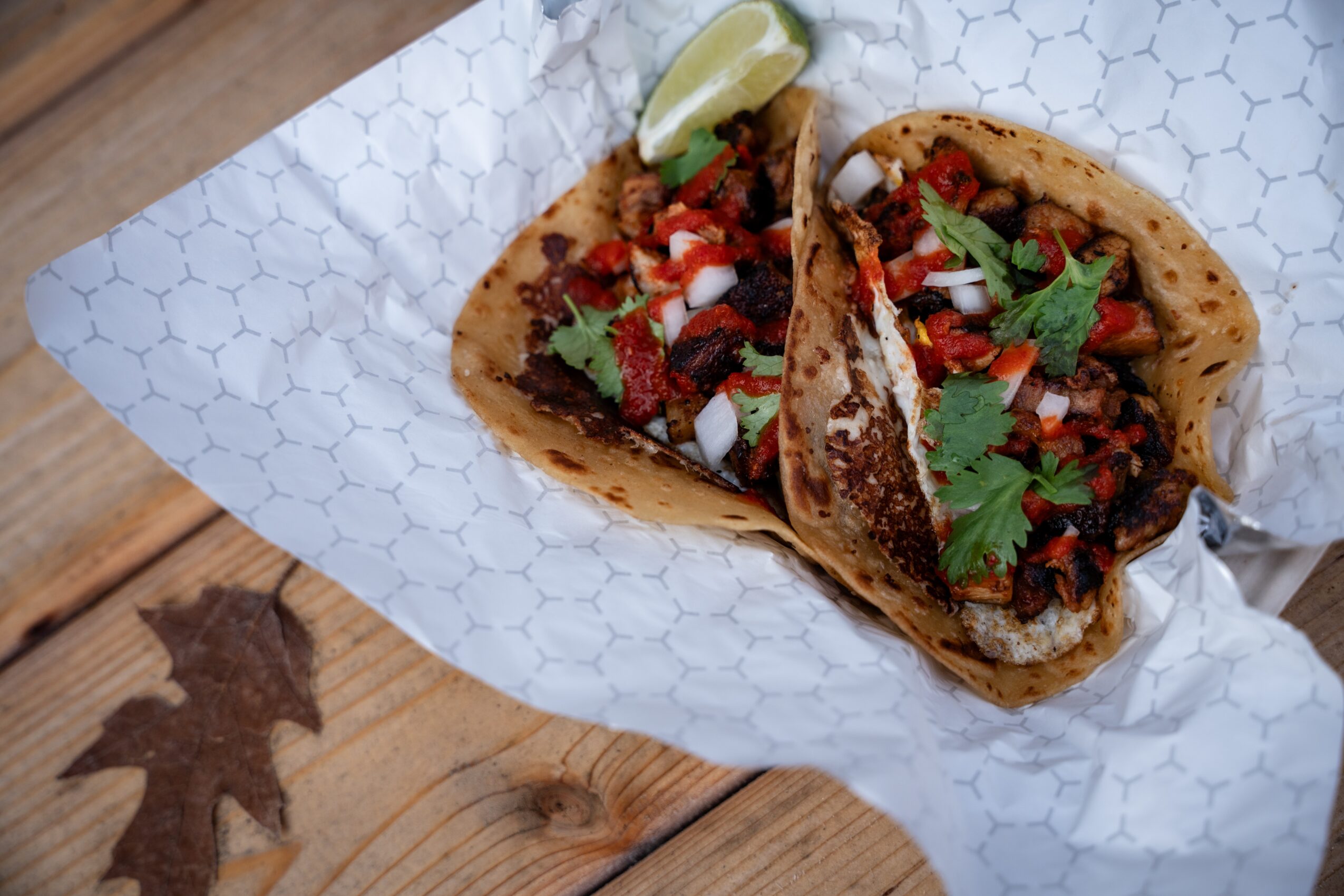 Two tacos with meat, vegetables, and cilantro on paper, with lemon wedge, on wooden surface.