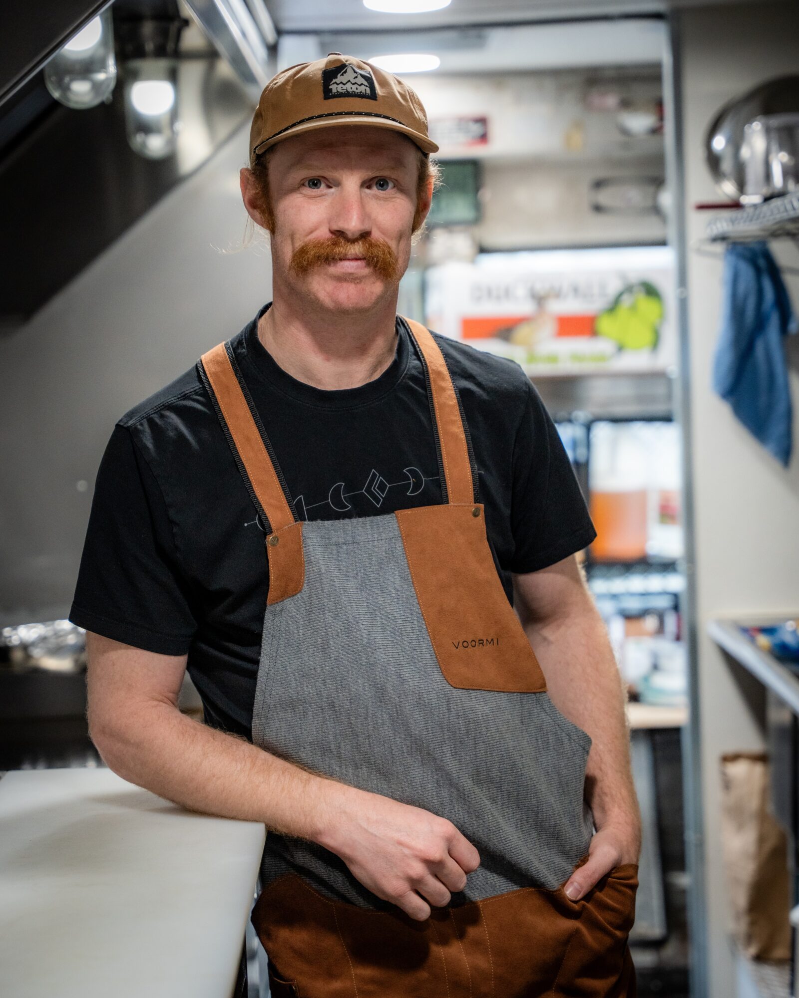 Man with a beard and mustache wearing a cap, apron, and black t-shirt, standing inside a kitchen or food truck.