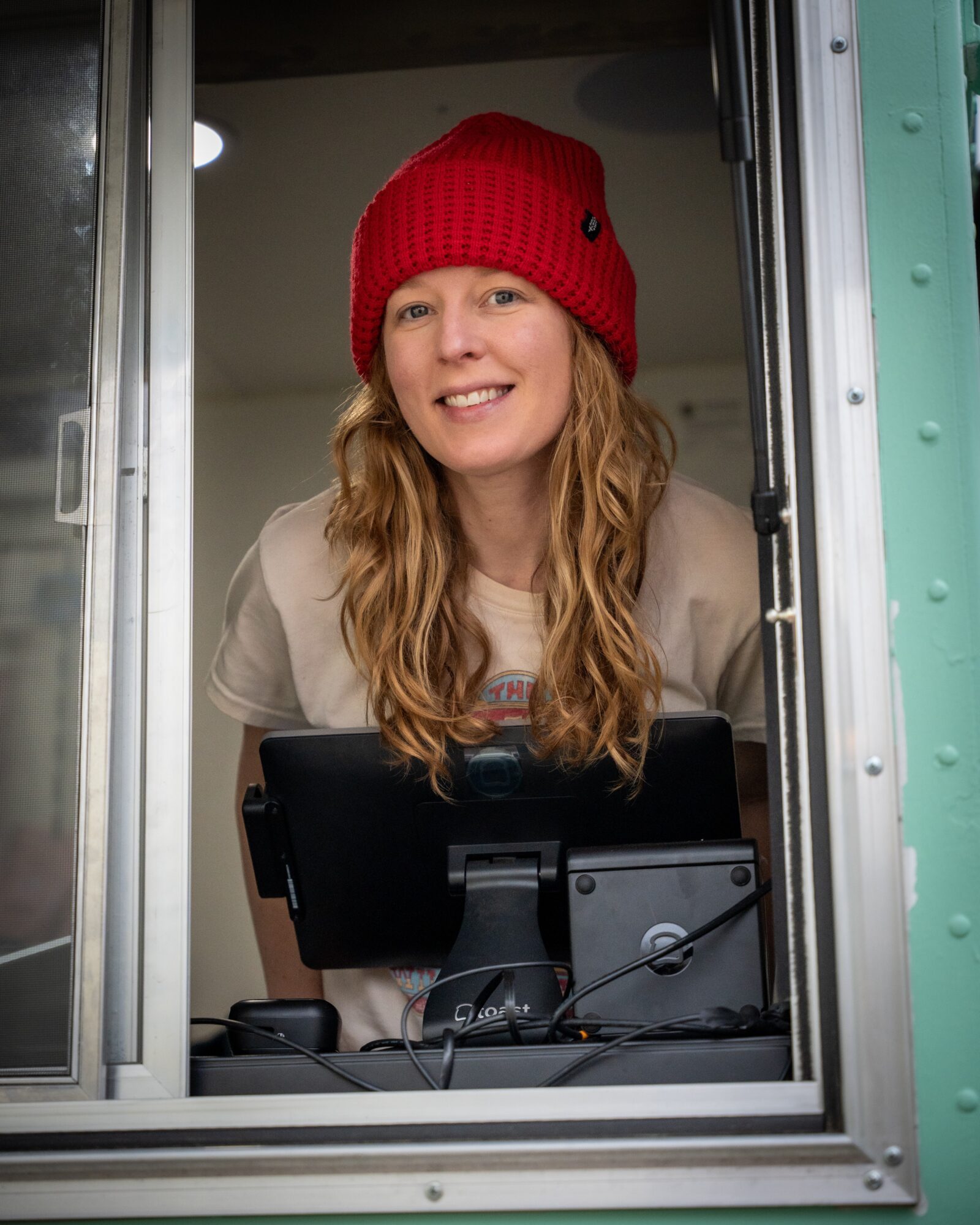 Woman with long hair wearing a red beanie, smiling, standing at a window with a computer, indoors.