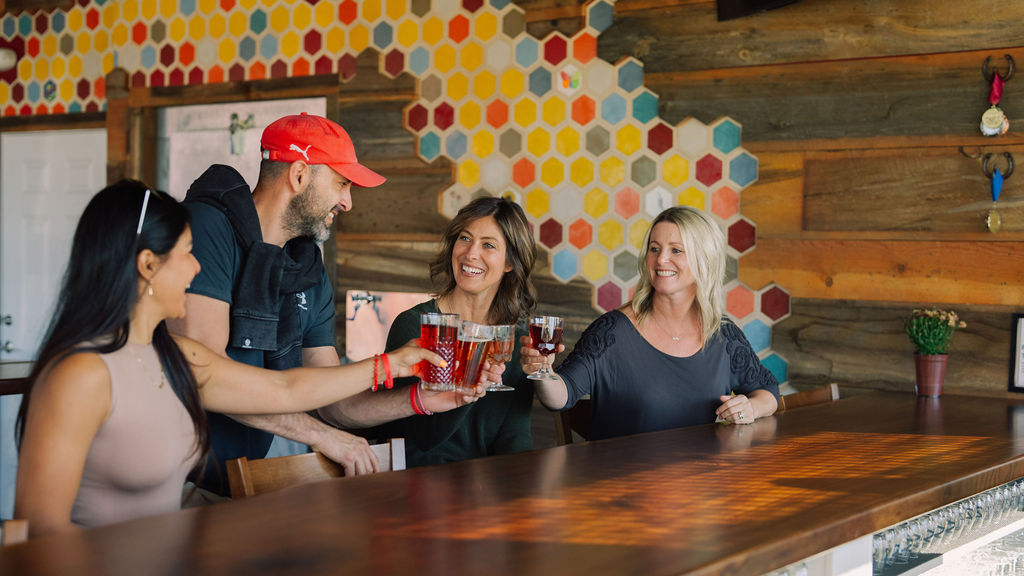 Four people sitting at a bar, clinking glasses, smiling, with colorful hexagon wall and wooden decor in background.